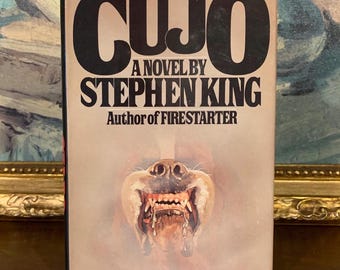 May include: A hardcover book titled Cujo, a novel by Stephen King, author of Firestarter. The cover features large black text on a light gray background, with a central illustration of a snarling dog showing its teeth and drooling saliva.