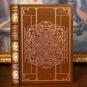Leather-bound edition of Dante's 'Divine Comedy' from The Franklin Library, featuring an ornate gold-embossed design on the cover with intricate patterns and symbols, displayed against a backdrop of a framed painting.