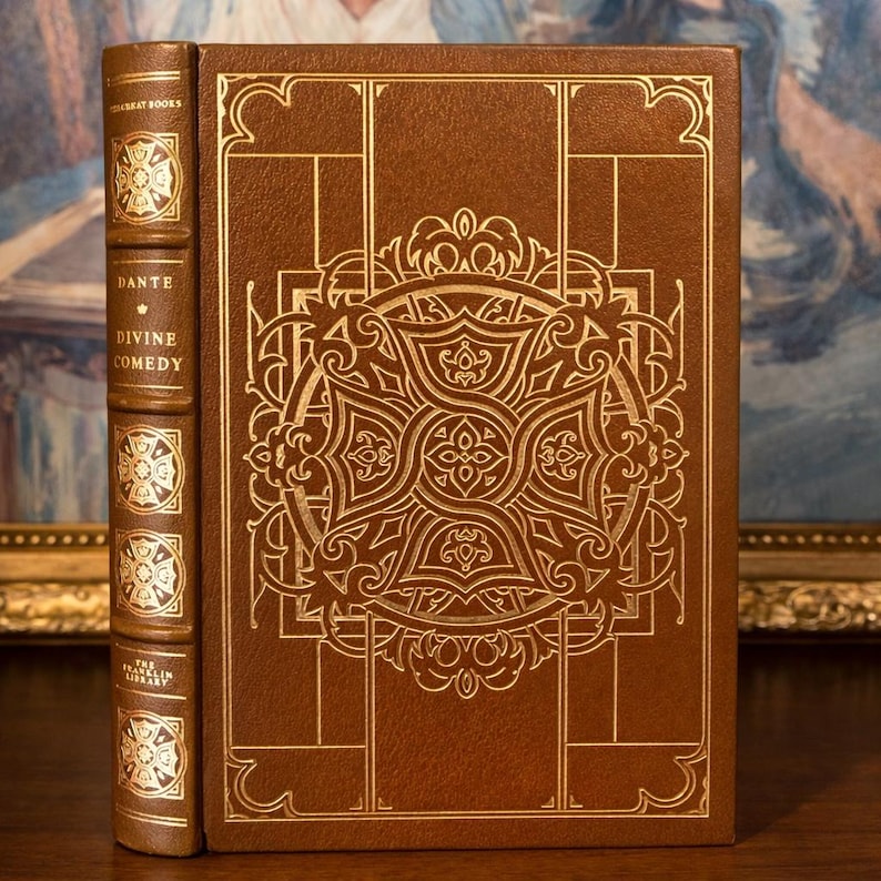 Leather-bound edition of Dante's 'Divine Comedy' from The Franklin Library, featuring an ornate gold-embossed design on the cover with intricate patterns and symbols, displayed against a backdrop of a framed painting.