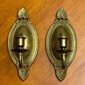 Puede incluir: Dos apliques de pared antiguos de latón. Cada aplique tiene un marco ovalado con un portavelas y una base decorativa. Los apliques son de latón y tienen un aspecto vintage. Los portavelas están diseñados para una sola vela.