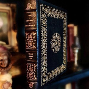 Vintage Leather-bound edition of “Tales of Mystery and Imagination” by Edgar Allan Poe with ornate gold detailing on the spine and cover. The book is displayed upright on a shelf next to antique objects, including a skull figure and vintage candleholder.