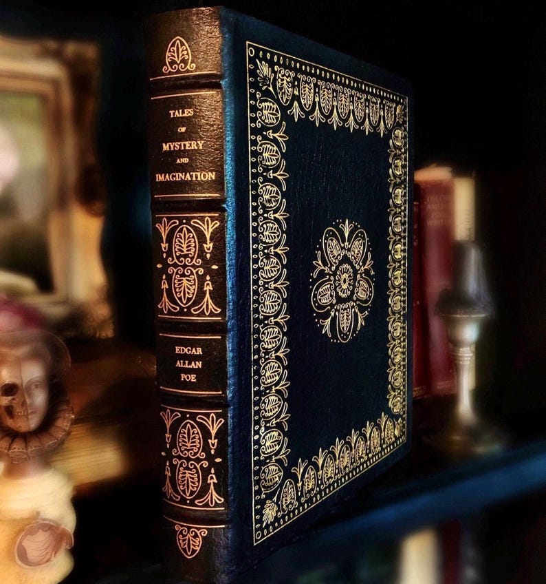 Vintage Leather-bound edition of “Tales of Mystery and Imagination” by Edgar Allan Poe with ornate gold detailing on the spine and cover. The book is displayed upright on a shelf next to antique objects, including a skull figure and vintage candleholder.