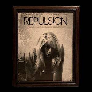 May include: A framed movie poster for the film "Repulsion" starring Catherine Deneuve and directed by Roman Polanski. The poster features a black and white photo of Deneuve looking directly at the viewer.