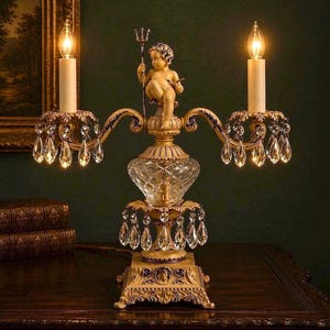 K&ouml;nnte beinhalten: Eine zweiarmige Kandelaber-Lampe mit einer zentralen Puttenfigur, die einen Dreizack h&auml;lt. Die Lampe hat einen goldfarbenen Metallsockel mit verzierten Details, ein Mittelst&uuml;ck aus klarem Schliffglas und zahlreiche h&auml;ngende Kristallprismen.