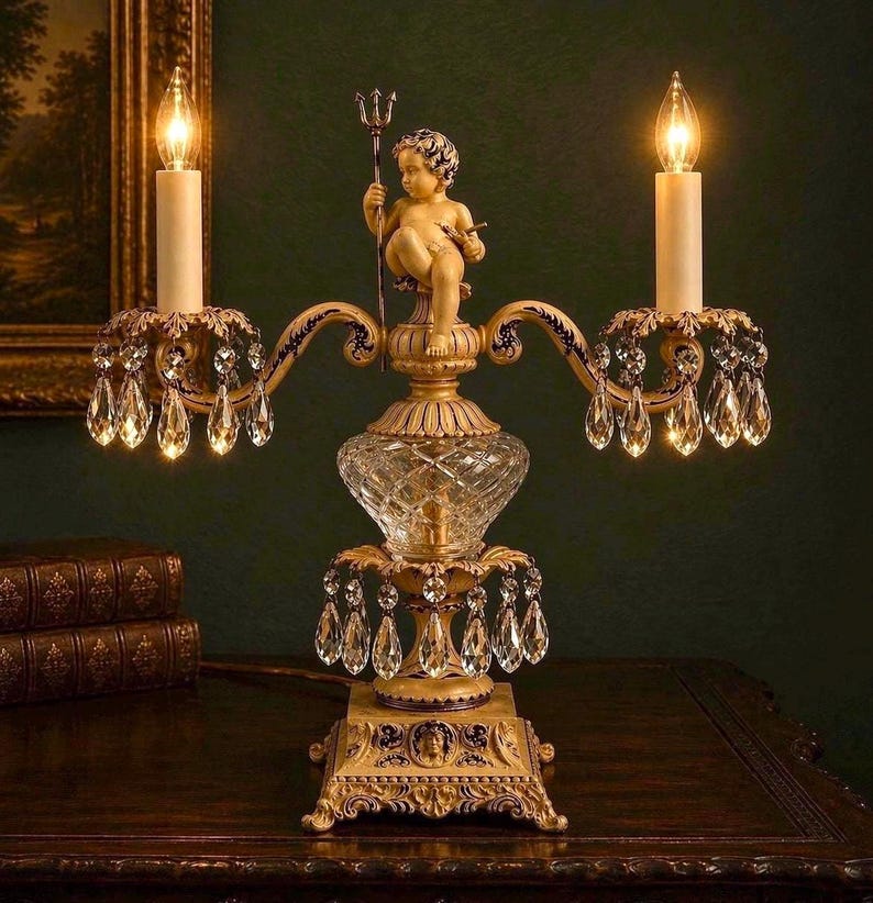 K&ouml;nnte beinhalten: Eine zweiarmige Kandelaber-Lampe mit einer zentralen Puttenfigur, die einen Dreizack h&auml;lt. Die Lampe hat einen goldfarbenen Metallsockel mit verzierten Details, ein Mittelst&uuml;ck aus klarem Schliffglas und zahlreiche h&auml;ngende Kristallprismen.