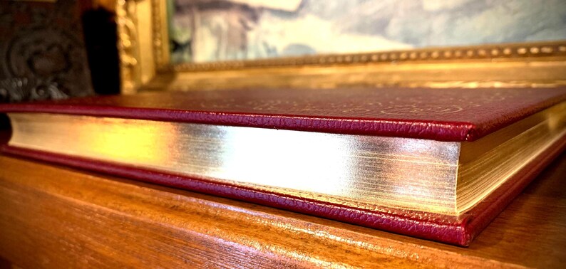 May include: A burgundy leather-bound book with gold-gilded edges. The book is closed and lying on a wooden surface.
