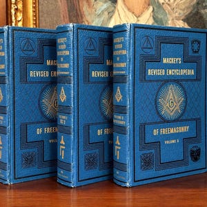 Puede incluir: Tres libros azules de tapa dura titulados "Mackey's Revised Encyclopedia of Freemasonry". Los libros tienen letras doradas y elementos decorativos. Los volúmenes 1, 2 y 3 son visibles. Los libros están colocados uno al lado del otro.