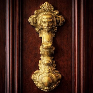May include: A detailed, close-up shot of a gold-colored door knocker. The knocker features a sculpted face at the top, with ornate scrollwork and a teardrop-shaped base. The knocker is mounted on a dark brown wooden door.