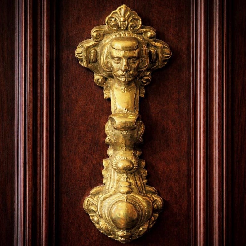 May include: A detailed, close-up shot of a gold-colored door knocker. The knocker features a sculpted face at the top, with ornate scrollwork and a teardrop-shaped base. The knocker is mounted on a dark brown wooden door.