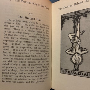 May include: Open book with text and an illustration of The Hanged Man tarot card. The card depicts a figure hanging upside down, with a halo, and the text "THE HANGED MAN" below. The text describes the card's symbolism.