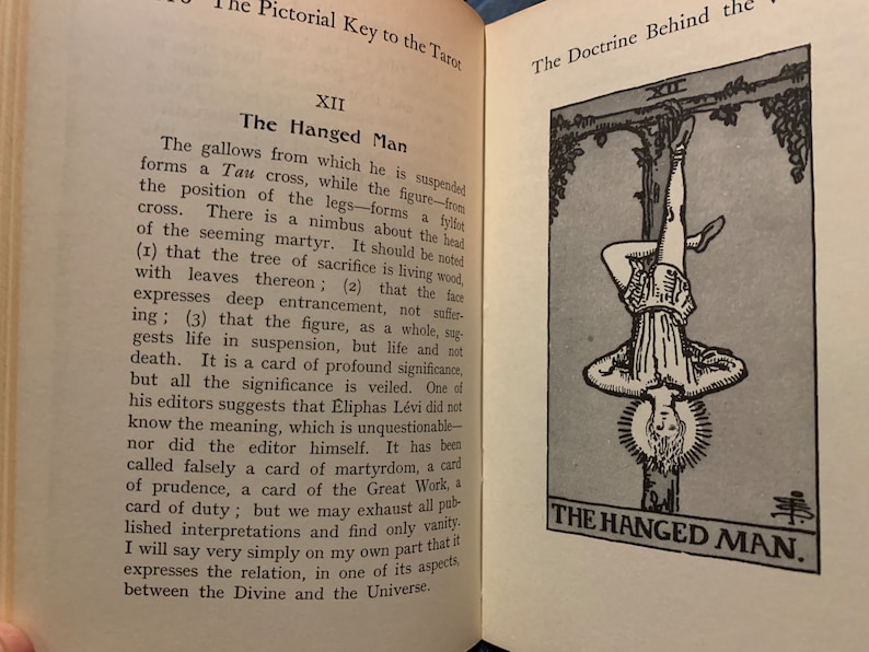 May include: Open book with text and an illustration of The Hanged Man tarot card. The card depicts a figure hanging upside down, with a halo, and the text "THE HANGED MAN" below. The text describes the card's symbolism.