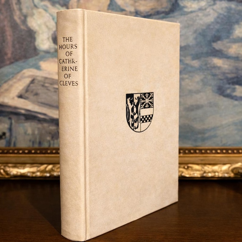 A book titled Hours of Catherine of Cleves, stand upright in a shelf, with a painting in the background.