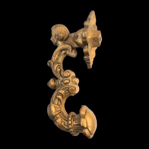 May include: Ornate, antique brass furniture hardware. The piece features a decorative design with a small cherub figure, scrolling details, and a curved handle. The hardware is likely for a drawer or cabinet.