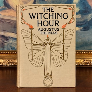 May include: A hardcover book titled The Witching Hour by Augustus Thomas with a beige cover. The front features a gold-ink illustration of a figure with large butterfly wings holding two torches, with a small clock face at the bottom.