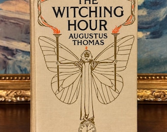 以下が含まれることがあります： オーガスタス・トーマス著『The Witching Hour』のベージュ色の表紙のハードカバー本。表紙には、大きな蝶の羽を持ち、2本の松明を掲げた人物の金色のインクのイラストと、下部に小さな時計の文字盤が描かれています。