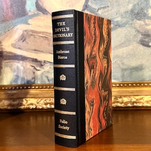 May include: A hardcover book titled "THE DEVIL'S DICTIONARY" by Ambrose Bierce. The spine is dark blue with gold lettering and decorative elements. The book's cover features a marbled pattern in shades of red, orange, and black.