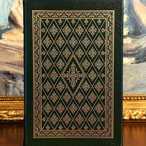 Vintage Leather-bound edition of The Poetry of Robert Frost in dark green with ornate gold gilt decoration on the cover, standing upright on a wooden surface in front of a framed painting.