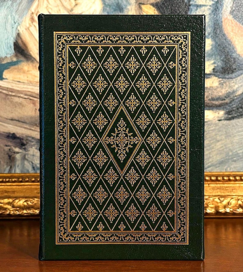Vintage Leather-bound edition of The Poetry of Robert Frost in dark green with ornate gold gilt decoration on the cover, standing upright on a wooden surface in front of a framed painting.