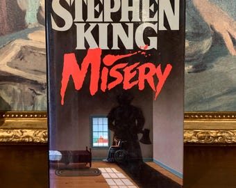 Puede incluir: Una portada de libro de bolsillo con un título rojo y negro "MISERY" de Stephen King. La portada presenta una silueta de una persona sosteniendo un hacha en una habitación con una cama y una persona en silla de ruedas.