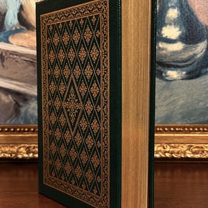 Vintage Leather-bound edition of The Poetry of Robert Frost in dark green with ornate gold gilt decoration on the cover, standing upright on a wooden surface in front of a framed painting.