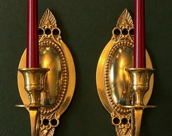 May include: A pair of matching gold-toned metal wall sconces, each featuring an oval backplate with decorative filigree detailing at the top and bottom. A single deep red taper candle is inserted into the central cup of each sconce.