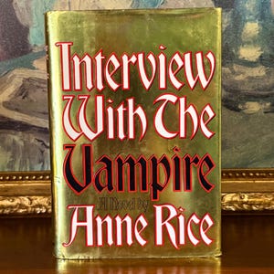 May include: A hardcover book titled "Interview With The Vampire" by Anne Rice. The book has a gold cover with the title in large, red and white lettering. The author's name is in white lettering at the bottom.