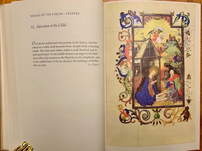 A medieval illuminated manuscript page with a depiction of the Nativity. The scene is set in a stable with a thatched roof. Mary kneels in a blue robe, holding the baby Jesus. Joseph stands behind her in a red robe. An angel with wings appears above the stable in a yellow sky. The text on the page reads 'HOURS OF THE VIRGIN-VESPERS 12. Adoration of the Child.