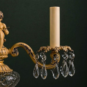 K&ouml;nnte beinhalten: Eine Wandleuchte mit einer cremefarbenen Puttenfigur, einem geschwungenen Arm mit schwarzen Schn&ouml;rkeldetails und einer zentralen Kerzenh&uuml;lse. Die Leuchte ist mit einer goldfarbenen blattf&ouml;rmigen Tropfschale und klaren Kristallprismen verziert.