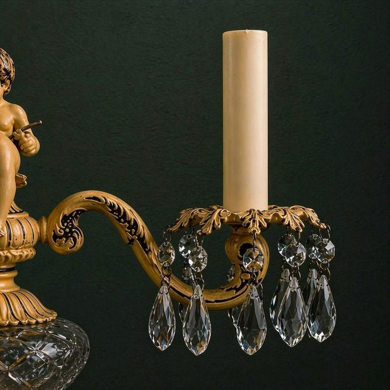 K&ouml;nnte beinhalten: Eine Wandleuchte mit einer cremefarbenen Puttenfigur, einem geschwungenen Arm mit schwarzen Schn&ouml;rkeldetails und einer zentralen Kerzenh&uuml;lse. Die Leuchte ist mit einer goldfarbenen blattf&ouml;rmigen Tropfschale und klaren Kristallprismen verziert.