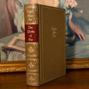 Può includere: Un libro d'epoca intitolato "The Works of Poe" con lettere dorate in rilievo e motivi decorativi sul dorso. Il libro ha un'etichetta bordeaux e una copertina verde tenue. Il libro è esposto su una superficie di legno.