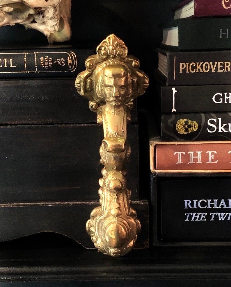 May include: A detailed, antique brass cabinet pull with a sculpted face and ornate design. The pull is vertically oriented, with a long handle and decorative elements. The background includes a stack of books with dark covers.