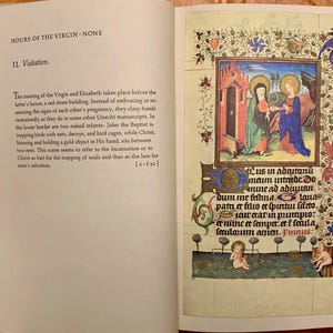An open book with a page showing a hand-drawn illustration of a scene from the Bible. The scene depicts the Visitation of the Virgin Mary to Elizabeth. The illustration is surrounded by a decorative border with floral motifs. The text on the page reads: 'Lus in adrutoni maim intende. Do mune ad adduian dum me fettina. Loua patu er filio er spurtun farto. laweratin punaριο: er nunc et semper er i feula lexulosum anien. Vinnus:'