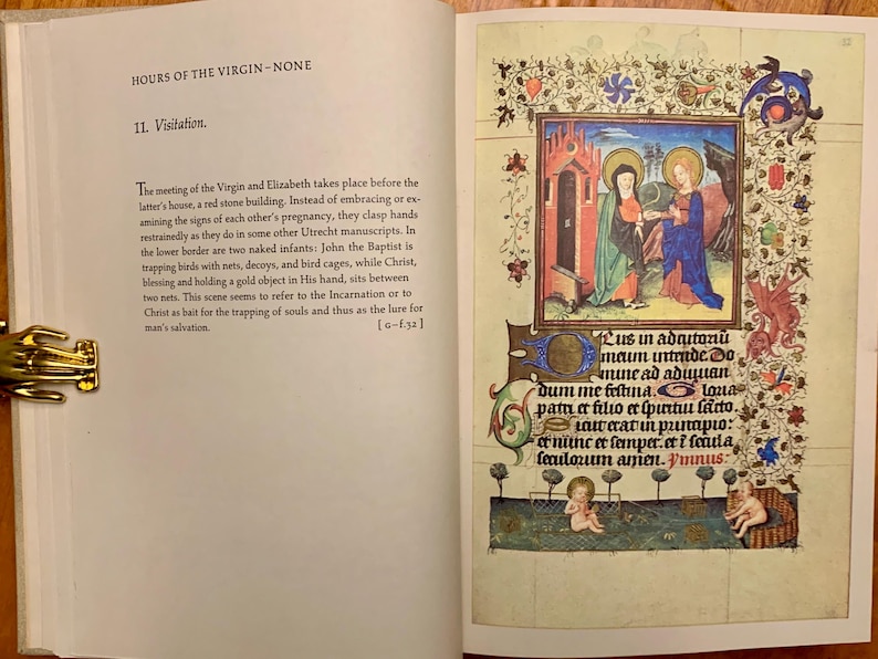 An open book with a page showing a hand-drawn illustration of a scene from the Bible. The scene depicts the Visitation of the Virgin Mary to Elizabeth. The illustration is surrounded by a decorative border with floral motifs. The text on the page reads: 'Lus in adrutoni maim intende. Do mune ad adduian dum me fettina. Loua patu er filio er spurtun farto. laweratin punaριο: er nunc et semper er i feula lexulosum anien. Vinnus:'
