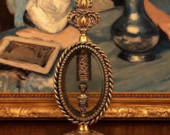 May include: A gold-toned metal decorative object featuring an oval frame with a twisted rope border. Inside the glass-enclosed oval, a small metallic bust of a person sits below a cylindrical, ornate, textured metal component. The piece has a decorative finial at the top and a wide, circular, engraved base.