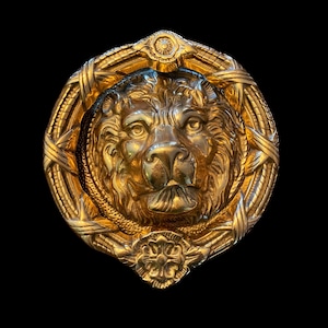 May include: A gold lion's head door knocker with a detailed design. The lion's head is surrounded by a wreath of leaves and flowers.