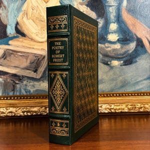 Vintage Leather-bound edition of The Poetry of Robert Frost in dark green with ornate gold gilt decoration on the cover, standing upright on a wooden surface in front of a framed painting.