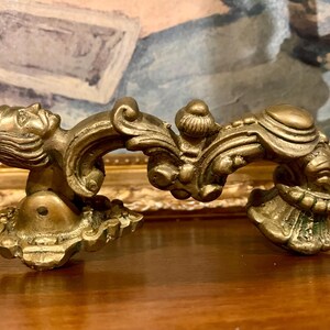 May include: Ornate brass furniture hardware with a detailed, sculpted design. The piece features a classical motif with a human face and scrolling embellishments. The hardware is likely for a drawer or cabinet, adding a touch of vintage elegance.