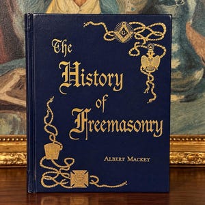 Peut inclure: Un livre relié bleu foncé intitulé The History of Freemasonry par Albert Mackey. La couverture présente des lettres dorées en relief et des motifs de corde dorés complexes avec des symboles maçonniques, dont une équerre et un compas.