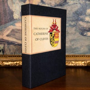 A vintage religious book inside it’s slipcase with the title, The Hours of Catherine of Cleves, stands upright on a shelf, with a gold framed painting in the background.