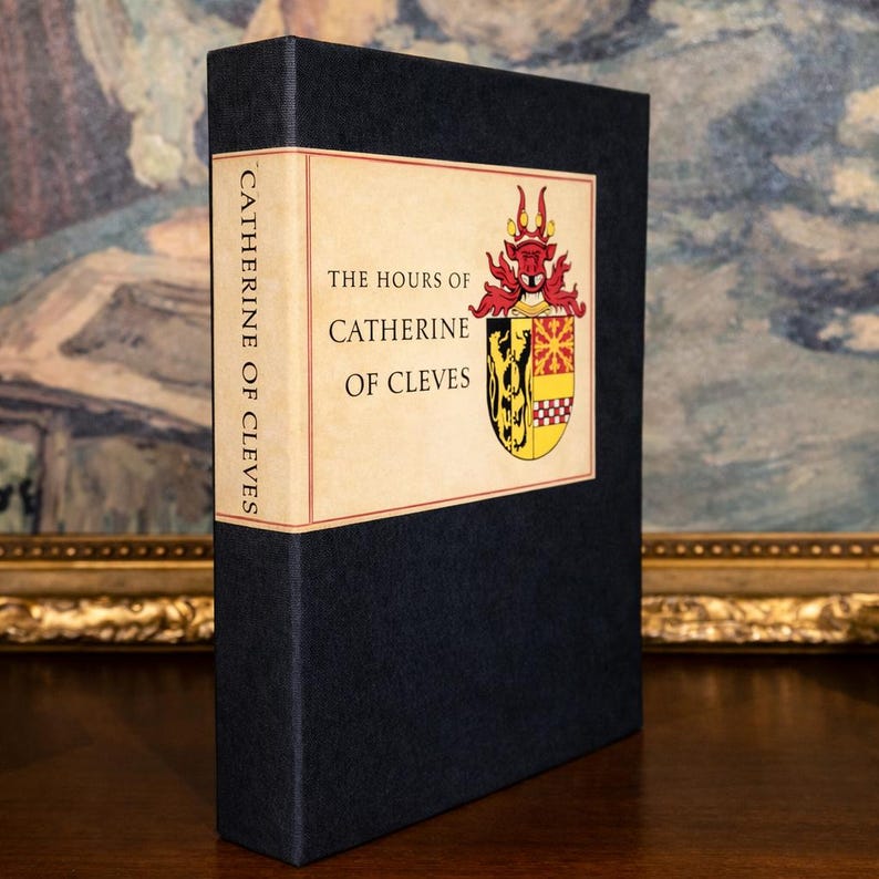 A vintage religious book inside it’s slipcase with the title, The Hours of Catherine of Cleves, stands upright on a shelf, with a gold framed painting in the background.