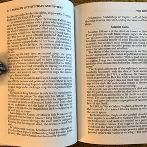 Puede incluir: Libro abierto sobre brujer&iacute;a y satanismo, sostenido por un sujetalibros met&aacute;lico en forma de mano. El texto trata sobre demonios, rituales y figuras hist&oacute;ricas relacionadas con el ocultismo.