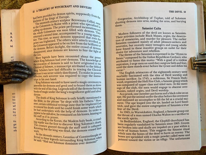 Puede incluir: Libro abierto sobre brujer&iacute;a y satanismo, sostenido por un sujetalibros met&aacute;lico en forma de mano. El texto trata sobre demonios, rituales y figuras hist&oacute;ricas relacionadas con el ocultismo.