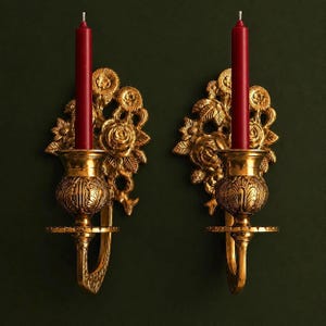 Puede incluir: Un par de apliques de pared de metal dorado con intrincados motivos florales y de hojas. Cada aplique sostiene una vela cónica de color rojo intenso. Los portavelas tienen una base texturizada y un brazo curvo en la placa posterior.