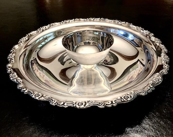 May include: A silver serving dish with a decorative rim and a smaller bowl in the center. The dish is designed for serving snacks or dips.