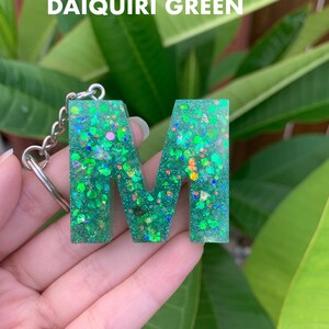 May include: A green glitter keychain with the letter M. The keychain is made of resin and has a silver chain. The background is a green plant.