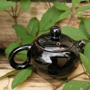 May include: A black ceramic teapot with a lid and a spout. The teapot is sitting on a wooden surface with green leaves.