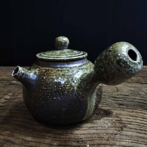 May include: A small, dark brown and green ceramic teapot with a unique spout and lid. The teapot has a rustic, handmade look.