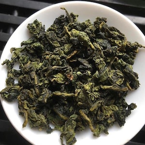 May include: Loose green tea leaves in a white bowl. The tea leaves are dried and ready to be brewed.