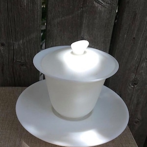 May include: White ceramic teacup and saucer set with a lid. The teacup has a rounded shape and a wide mouth. The saucer is flat and has a slightly raised edge. The lid has a small knob on top.