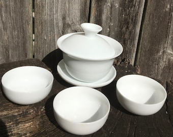 Könnte beinhalten: Ein weißes Keramik-Tee-Set mit einem Teekännchen mit Deckel, einer Untertasse und drei kleinen Teetassen. Das Set ist auf einer Holzoberfläche angeordnet.